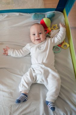 Mutlu bir bebek kolları havada, oyun parkında sırtüstü yatarken gülümser. Kafasının yanında renkli peluş ve plastik oyuncaklar var..