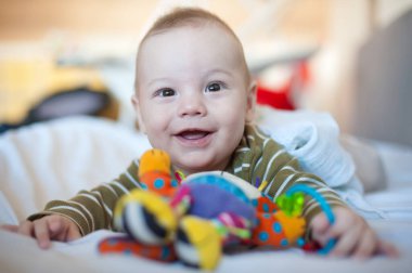 Yumuşak oyuncaklarla çevrili bir yatakta bir bebek yatıyor. Bebek parlak, renkli oyuncaklara uzanırken gülümsüyor. Güneş ışığı odayı dolduruyor..