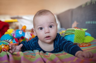 Oturma odasında battaniyenin üzerinde bir bebek yatıyor. Bebek çeşitli renkli oyuncaklarla çevriliyken şaşırmış ve meraklı görünüyor. Odada güneş ışığı parlıyor..
