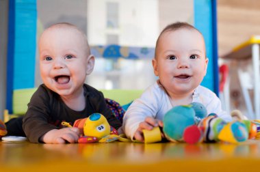 İkiz bebekler renkli bir paspasın üzerinde karınlarının üzerinde yatarlar. Çeşitli yumuşak oyuncaklarla oynarken gülümserler. Odanın parlak renkleri ve yeterli ışığı var..