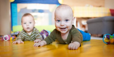 Parlak bir oyun alanında renkli bir paspasın üzerinde iki bebek emekliyor. Bir bebek merakla bakarken diğeri gülümsüyor. Etraflarına oyuncaklar saçılmış..