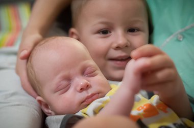 Neşeli bir ev sahnesinde, bir ağabey gülümserken küçük kardeşini nazikçe kucağına alır. Aile hayatının yumuşak arka planına karşı bağları parlıyor..