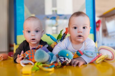 İki ikiz kardeş bebek, yumuşak oyuncaklarla çevrili canlı bir oyun hasırının üzerinde yatıyor. Yakınlardaki renkli nesnelerle etkileşime girerken merak ve meraklarını dile getiriyorlar..