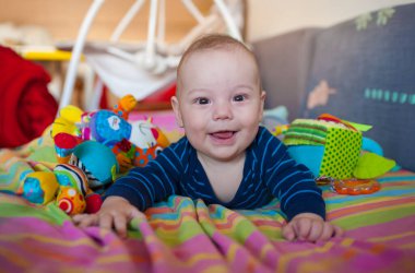 Bir bebek yumuşak bir battaniyeye uzanır ve renkli oyuncaklara uzanırken gülümser. Oda aydınlık ve davetkar, bebeğin etrafında eğlenceli şeylerle dolu..