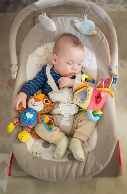 Bebek, yumuşak oyuncaklarla çevrili bir fedainin içinde huzur içinde uyuyor. Oda aydınlık, ve bebek rahat giysiler içinde dinleniyor, sessiz bir anın tadını çıkarıyor..