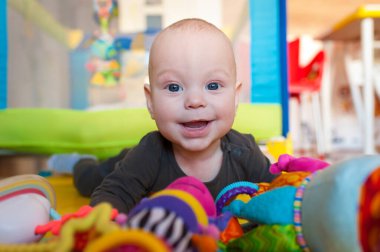 Neşeli bir bebek, neşeli bir oyun hasırının üzerinde uzanırken, güneşli, davetkar bir odada sıcaklık ve neşe saçan pelüş oyuncaklarla çevrili bir şekilde sırıtır..