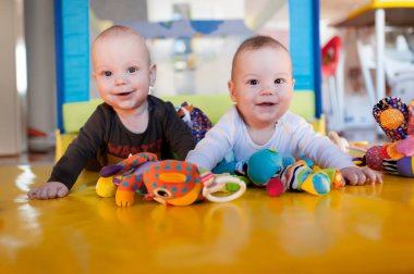 İki neşeli bebek bir dizi renkli oyuncakla çevrili canlı sarı bir hasırda birlikte oynuyorlar. Gülüşleri ve kahkahaları rahat oyun odasında neşeli bir atmosfer yaratıyor..
