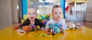 İki sevimli ikiz renkli oyuncaklarla çevrili canlı sarı bir masada oturuyorlar. Neşe ve merak ifadeleri, güneş ışığı içeri girerken sıcacık mekanı aydınlatıyor..