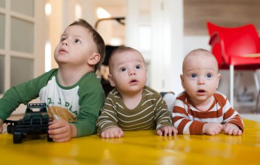 Üç kardeş evdeki parlak sarı oyun minderinde merak ve merak dolu bir anı paylaşıyorlar. Büyük kardeş oyuncakla oynarken meraklı ikizler etrafa bakınıyor, merak ve heyecan dolu..