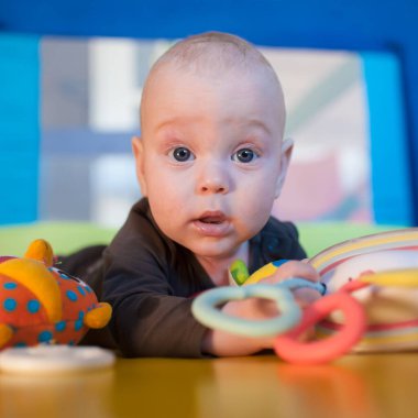 Meraklı bir bebek renkli oyuncaklarla çevrili canlı bir oyun hasırının üzerinde oturur. Geniş gözlerle uzanır ve evde oyun saatinde neşeli bir ifade takınır..