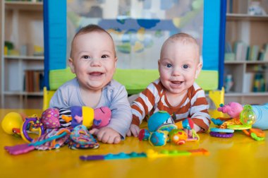 İki mutlu ikiz bebek renkli oyuncaklarla çevrili canlı bir masada oturuyor. Gülüşleri odayı aydınlatıyor, kahkahalarla dolu neşeli bir atmosfer yaratıyor..
