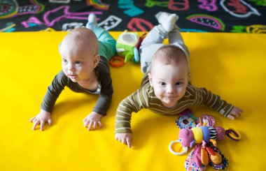 İki küçük kardeş sarı bir hasırın üzerinde sürünüyorlar. Önlerine konulan oyuncaklara uzanıyorlar. Odanın zemininde çizimler ve tasarımlar olan renkli bir zemin var..