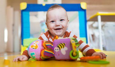 Sıcak ve davetkar bir oyun alanında, parlak gözlü mutlu bir bebek canlı oyuncaklara uzanırken geniş bir gülümseme sergiliyor. Yumuşak paspaslar ve neşeli bir ortam oluşturur..