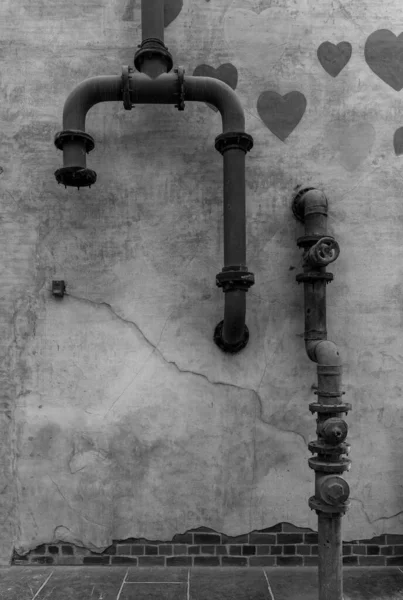 A black and white picture of a partially broken wall with hearts drawn and with some exposed thick water pipes