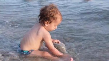 Küçük çocuk çakıl taşının üzerinde oturuyor ve suya taşlar atıyor. Rusya