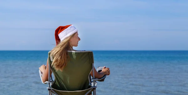 海のそばの大晦日のビーチを旅する クリスマスの帽子の女の子は太陽の下で日光浴をしています カメラに背を向けて椅子に座っている女性が遠くを見ている 水のそばにいる観光客 — ストック写真