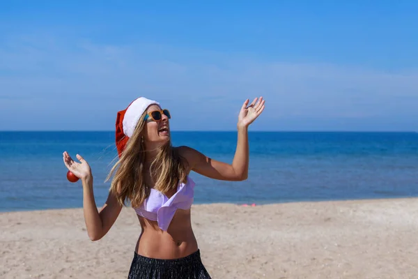 ビーチでの新年 手にクリスマスツリーのおもちゃでサンタの帽子の面白い女の子 休日の装飾 — ストック写真