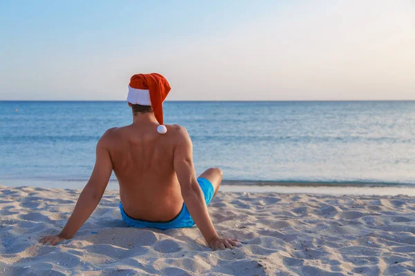 海のそばのビーチでの大晦日 クリスマスの帽子の男は太陽の下で日光浴をしている 青い空をコピースペースのように — ストック写真