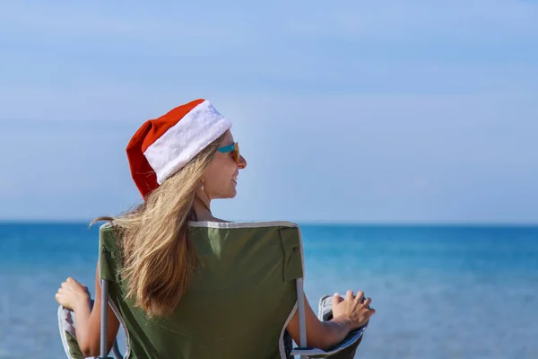 海のそばのビーチで大晦日に旅行します クリスマスの帽子の女の子は日光浴です 暗い眼鏡をかけた女だ カメラに背を向けて椅子に座っている女性は距離を見ています 水で休める観光 — ストック写真