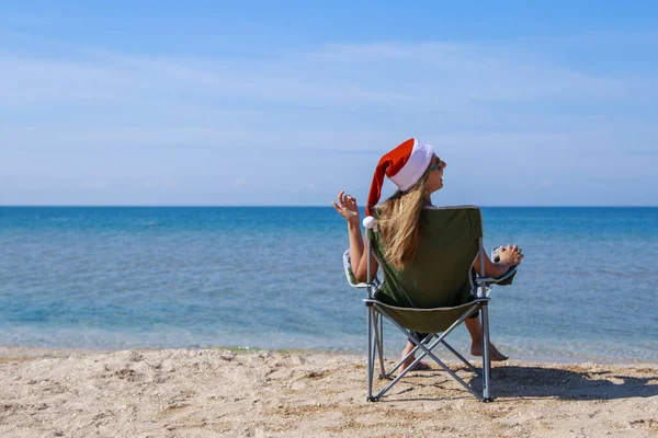 海のそばのビーチで大晦日に旅行します クリスマスの帽子の女の子は日光浴です 暗い眼鏡をかけた女だ カメラに背を向けて椅子に座っている女性は距離を見ています 水で休める観光 — ストック写真