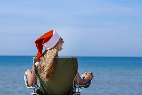 海のそばのビーチで大晦日に旅行します クリスマスの帽子の女の子は日光浴です 暗い眼鏡をかけた女だ カメラに背を向けて椅子に座っている女性は距離を見ています 水で休める観光 — ストック写真