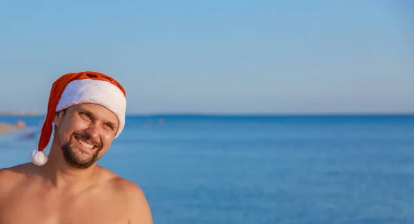 海のそばのビーチでの大晦日 クリスマスの帽子の男は太陽の下で日光浴をしている 青い空をコピースペースのように — ストック写真