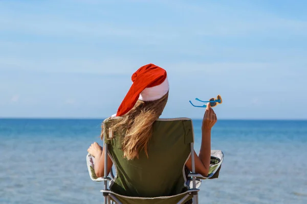 travel on New year\'s eve on beach by sea. girl in Christmas hat is sunbathing in sun. woma,n with dark glasses. woman in chair with her back to camera looks into distance. Tourist resting by water.