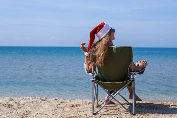 海のそばのビーチで大晦日に旅行します クリスマスの帽子の女の子は日光浴です 暗い眼鏡をかけた女だ カメラに背を向けて椅子に座っている女性は距離を見ています 水で休める観光 — ストック写真