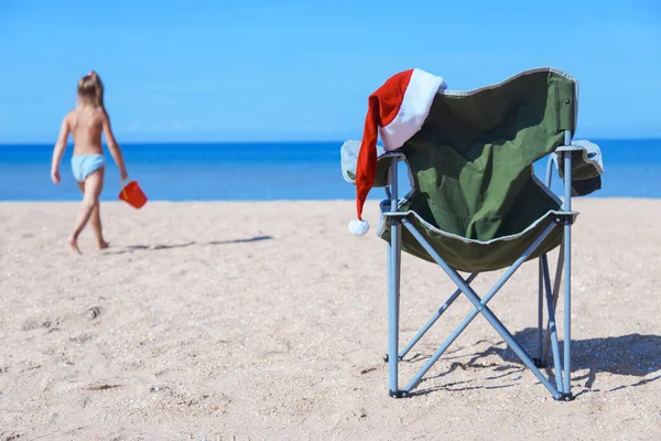 海沿いのビーチでの新年の前夜 観光客の椅子にサンタクロースの帽子 クリスマス休暇だ 熱い国での観光 背景に赤いバケツを持った女の子が砂の上を歩き — ストック写真