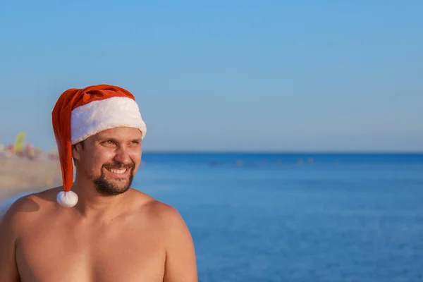 海のそばのビーチでの大晦日 クリスマスの帽子の男は太陽の下で日光浴をしている 青い空をコピースペースのように — ストック写真