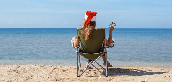 海のそばのビーチで大晦日に旅行します クリスマスの帽子の女の子は日光浴です 暗い眼鏡をかけた女だ カメラに背を向けて椅子に座っている女性は距離を見ています 水で休める観光 — ストック写真