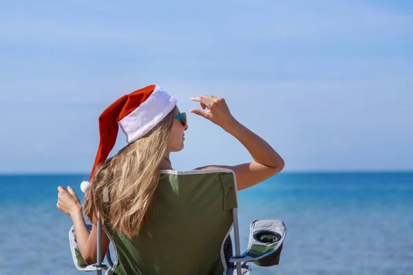 海のそばのビーチで大晦日に旅行します クリスマスの帽子の女の子は日光浴です 暗い眼鏡をかけた女だ カメラに背を向けて椅子に座っている女性は距離を見ています 水で休める観光 — ストック写真