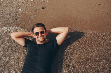 Tatildeki adam güneşlenir, sandalyede oturur, güneş gözlüğünü çıkarır ve gülümser.