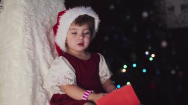 Kırmızı elbiseli bir çocuk yeni yılı bekliyor. Noel 'i gece yarısı kutlama konsepti. tatil kostümü. Bir kız Noel Baba 'ya mektup yazıyor. istek listesi