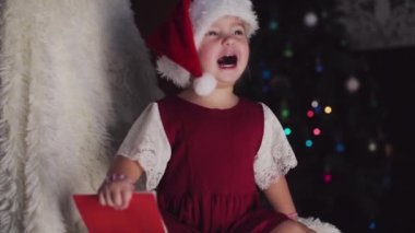 Kırmızı elbiseli bir çocuk yeni yılı bekliyor. Noel 'i gece yarısı kutlama konsepti. tatil kostümü. Bir kız Noel Baba 'ya mektup yazıyor. istek listesi