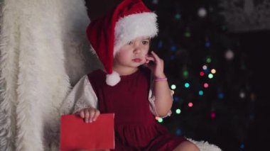 Kırmızı elbiseli bir çocuk yeni yılı bekliyor. Noel 'i gece yarısı kutlama konsepti. tatil kostümü. Bir kız Noel Baba 'ya mektup yazıyor. istek listesi