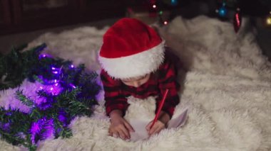 Kırmızı elbiseli bir çocuk yeni yılı bekliyor. Noel 'i gece yarısı kutlama konsepti. tatil kostümü. Bir kız Noel Baba 'ya mektup yazıyor. istek listesi