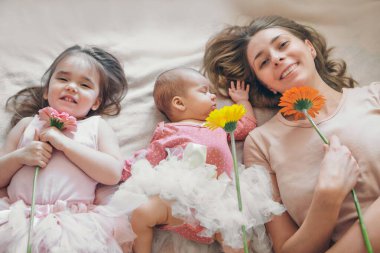 Çiçekli üç kızın portresi. Uyuyan yeni doğmuş bir bebek, küçük bir kız ve bir kadın yerde yatıyor. İç dekorasyon olarak çiçekler. 8 Mart