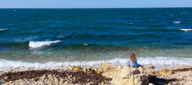Hava gözlemi konsepti, meteoroloji. Rüzgar ve küçük dalgalar. Kız kıyıda oturur ve uzaklığa bakar. kayalık kıyı