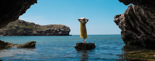 Una mujer con un vestido blanco y un sombrero negro de pie sobre las rocas junto al mar —  Fotos de Stock