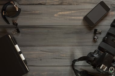 Ahşap arka planda fotoğrafçı için çeşitli nesneler. Akıllı telefon, kamera lensi, kol saati, fosforlu kalem, defter, klasörler.