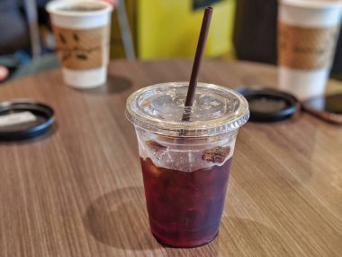 Buzlu, plastik bir fincanda samanla soğuk bir kahve, arka planda diğer kahve içecekleri olan rahat bir kafedeki ahşap bir masada oturmak.