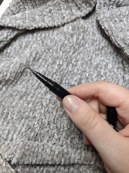 Hand holding a black eyeliner pen with the cap removed in front of a soft grey background