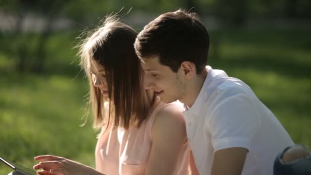 Der Kerl mit dem Mädchen, das im Park sitzt und auf das Tablet schaut. Nahaufnahme. — Stockvideo