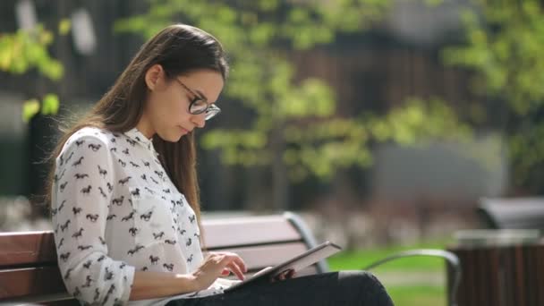 Hermoso estudiante en gafas con una tableta digital sentado al aire libre — Vídeo de stock