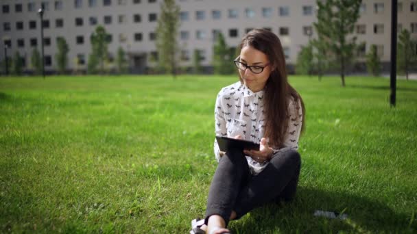 Gyönyörű diák szemüveg, digitális tabletta ül a fűben — Stock videók