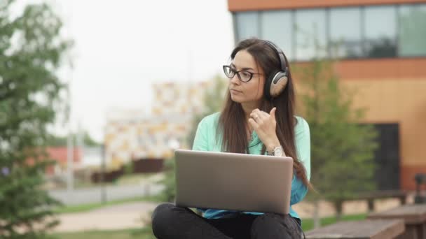 Attractive young girl with headphones sitting in the open space with a laptop. — Stock Video