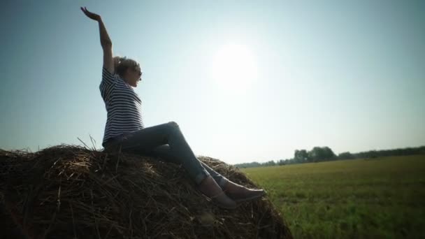 Pretty girl is sitting on haystacks and pulls hands to the sky. — Stock Video