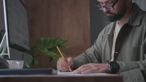 Een man werkt aan een project in zijn kantoor, schrijft en tekent — Stockvideo