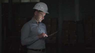 Kasklı ve gömlekli bir adam karanlık bir deponun önünde tablet kullanıyor.